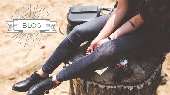 Kobieta siedzi na pieńku, jest w dżinsach, a obok jej nogi stoi puszka cooca coli i skórzana torebka   