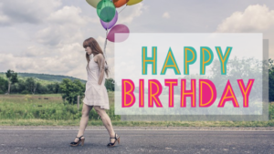 Kobieta przechodzi drogę, w ręku trzyma kolorowe balony. Napis ozdobny "Happy birthday"