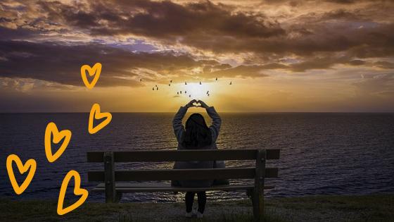 kobieta siedząca na ławce z wyciągniętymi ramionami nad głową, jej dłonie układają się w kształt serca. W tle zachód słońca na plaży.