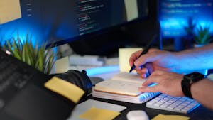 A person planning a software project with handwritten notes and code on a monitor in a modern workspace.